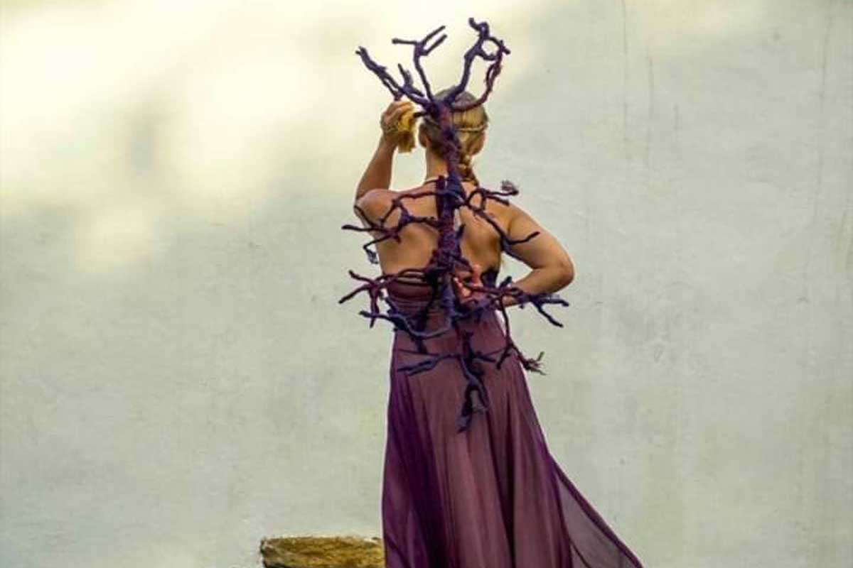 Uma pessoa está de costas na foto, caminhando em uma estrutura de prédio colonial, vestida com roupa de tecido roxo e ornamento que relembra um galho de árvore também roxo.
