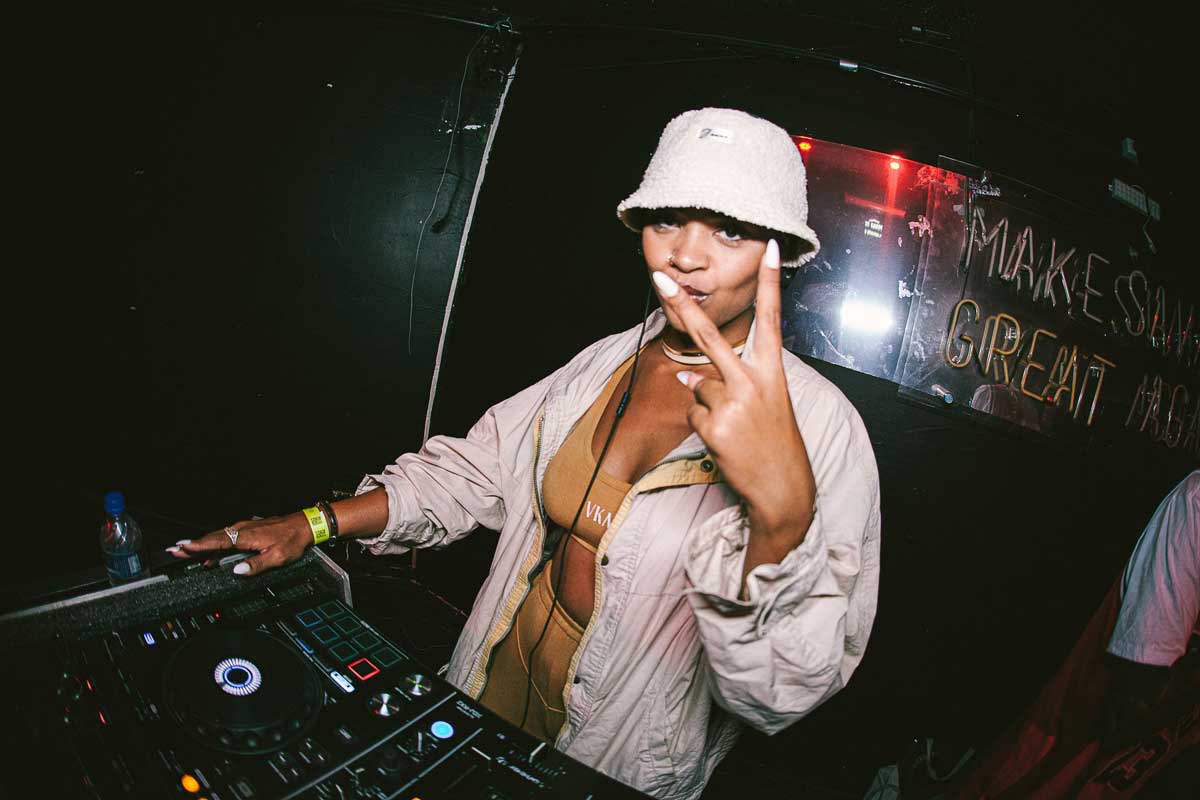 A foto mostra uma mulher operando uma mesa de DJ. Uma das mãos está apoiada na mesa e a outra mostra dois dedos levantados. A artista usa um conjunto de top e calça beje e uma jaqueta creme, além de chapéu, fone de ouvido e um colar. 