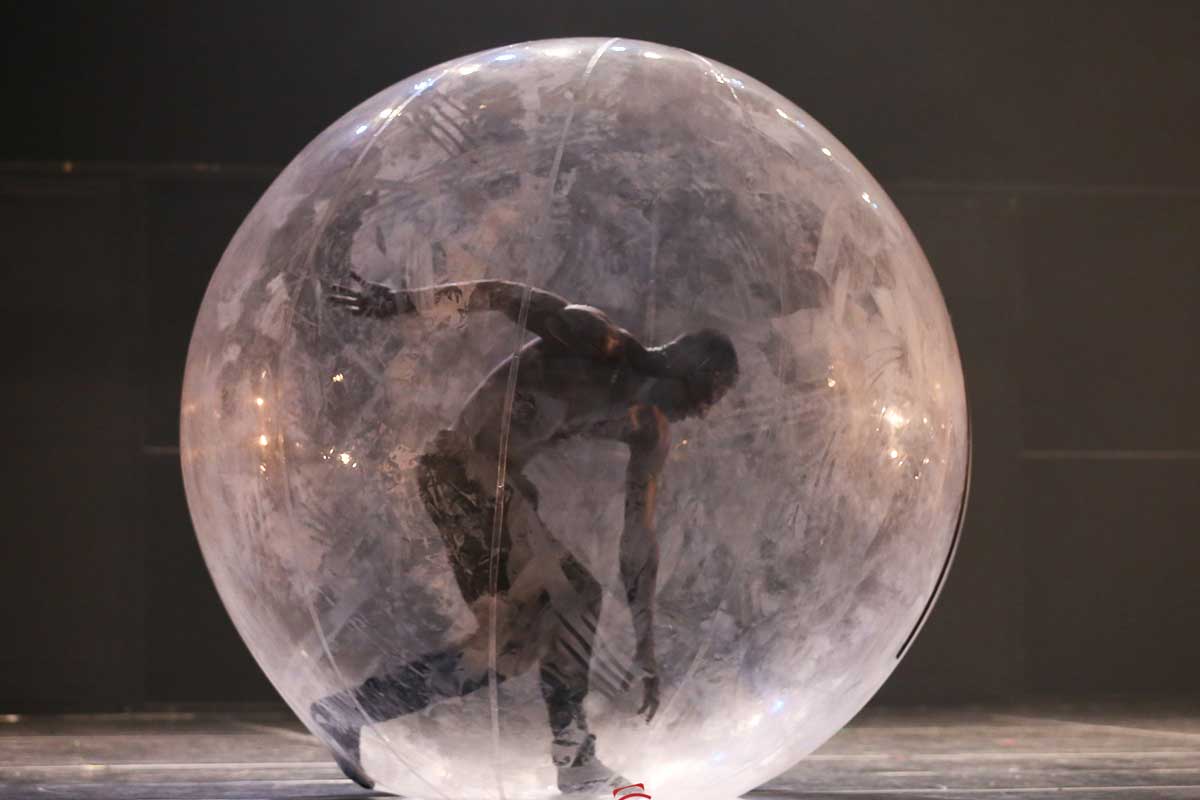 A foto mostra um homem, sem blusa e de calça, dentro de uma bolha de plástico transparente. O homem está em pé, em movimento e com o rosto virado para baixo. A bolha está sob um chão liso e fundo escuro.