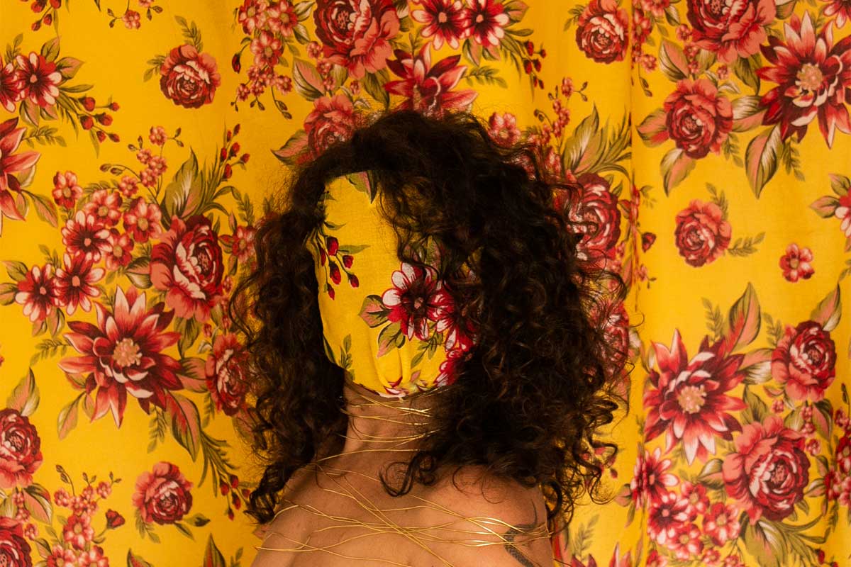 A imagem é uma foto com fundo de tecido amarelo com flores vermelhas, tem uma mulher centralizada, com cabelos ondulados escuros, rosto coberto pelo mesmo tecido que aparece ao fundo e sem blusa. A foto mostra dos ombros para cima e tem fios dourados pelo pescoço e ombros da mulher