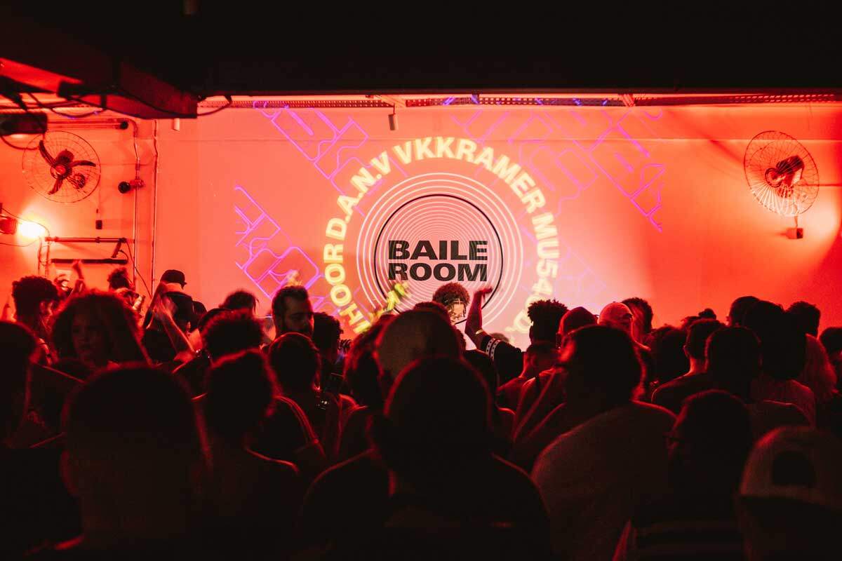 A foto mostra uma apresentação do artista do ponto de vista da plateia. As pessoas estão dançando enquanto o DJ toca em um ambiente fechado com luzes vermelhas. Ao fundo, na parede, há uma projeção com o nome “baile room”.