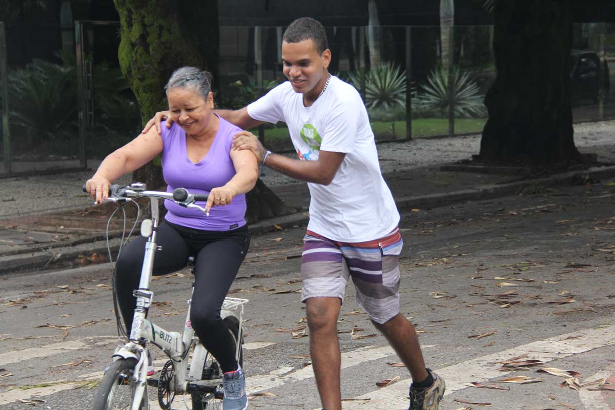 A foto mostra uma mulher aprendendo a andar de bicicleta na rua, guiada por um homem. Ela usa blusa roxa e calça preta e está em uma bicicleta branca. O homem usa blusa branca e bermuda listrada em tons de cinza, azul, branco e vermelho.