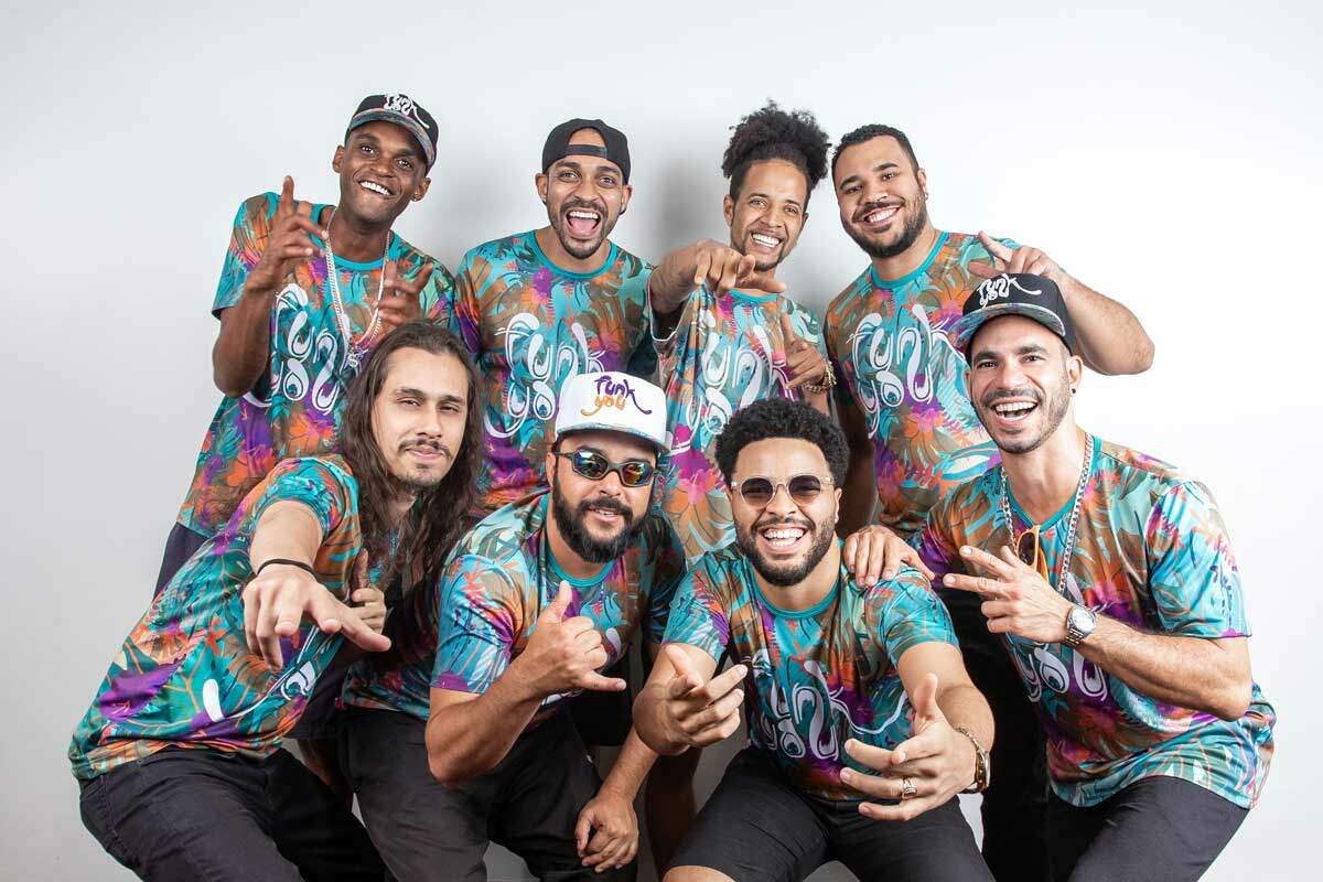 A foto mostra oito homens sorrindo e fazendo gestos com as mãos e usando camisas coloridas com o escrito “funk you”. Quatro deles estão agachados na frente e os outros quatro estão em pé, atrás. 