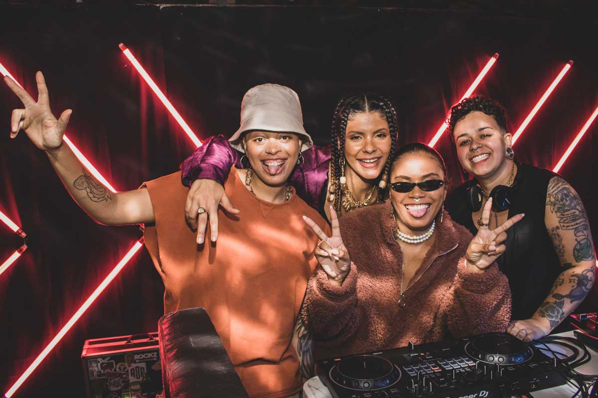 A imagem é uma fotografia de quatro DJ`s mulheres, em cima de um palco, atrás de uma mesa controladora de som. As quatros mulheres estão próximas umas das outras e todas estão sorrindo e olhando para câmera. O fundo é escuro com lâmpadas retangulares finas vermelhas de led. 