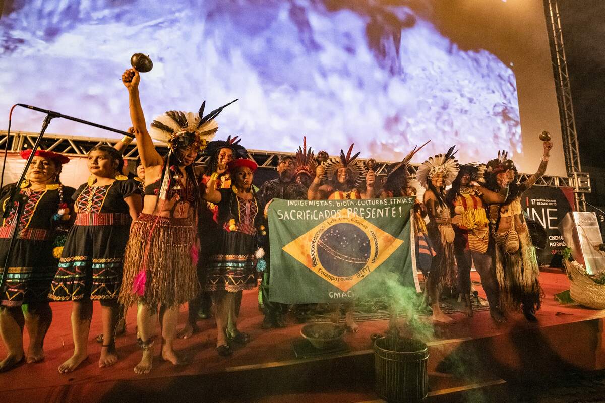 A imagem é uma fotografia de um grupo de indígenas, em pé, lado a lado, em cima de um palco, com um telão ao fundo. Os e as indígenas estão com os braços erguidos, segurando chocalhos e uma bandeira do Brasil, onde lê-se "indigenistas, biota, culturas, etnias… sacrificados. Presente! CMACI"