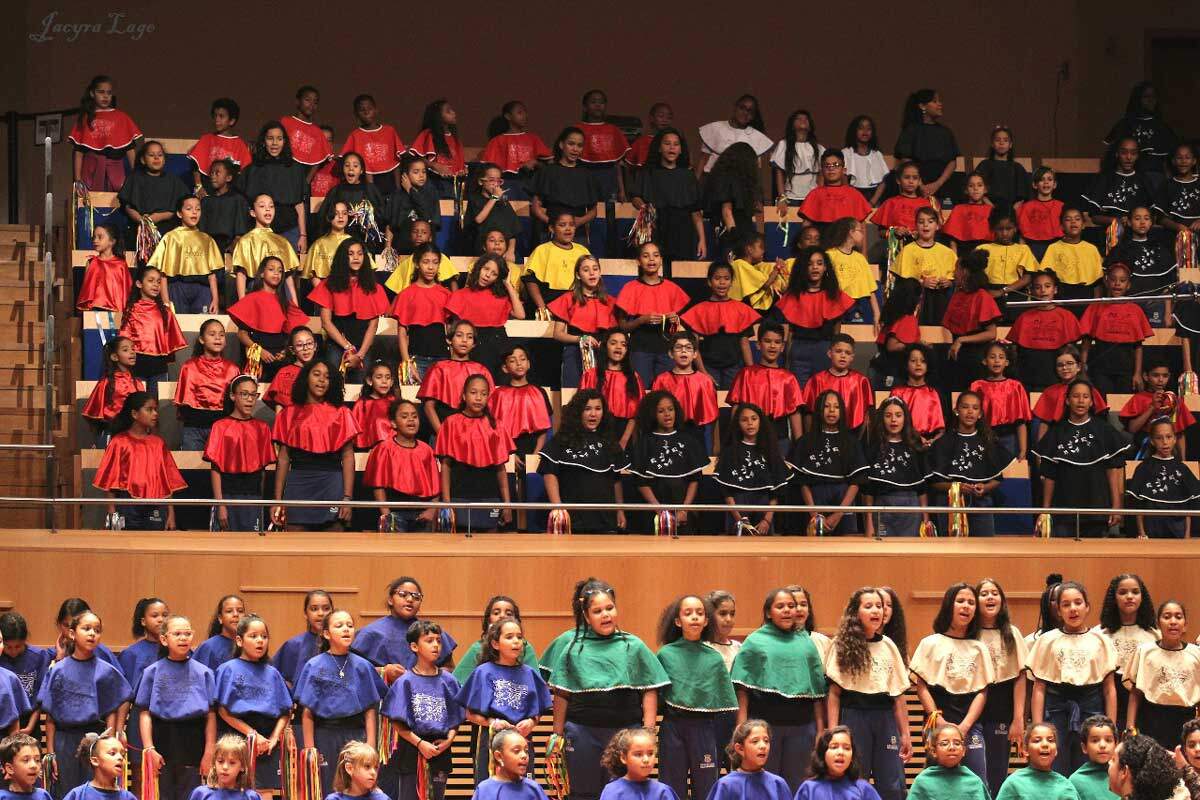 A foto mostra jovens cantando em um coral. Há dezenas deles e estão posicionados em níveis diferentes, como em uma escada. Eles usam túnicas com cores variadas, como vermelho, amarelo, azul, verde, branco e bege.
