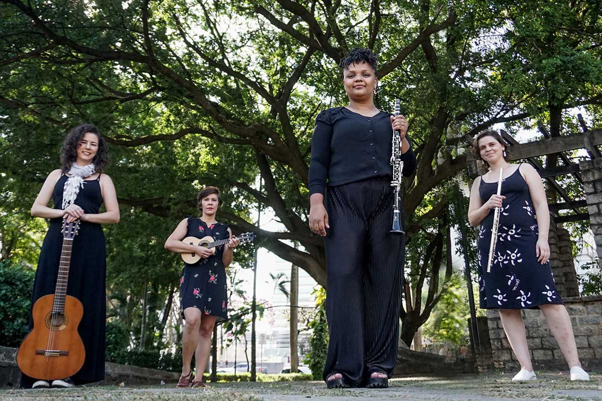 A imagem é uma fotografia de quatro mulheres em uma praça com árvores ao fundo, todas vestidas de preto e em pé. Todas estão olhando para câmera e seguram um instrumento musical, quais sejam violão, cavaquinho, clarinete e flauta