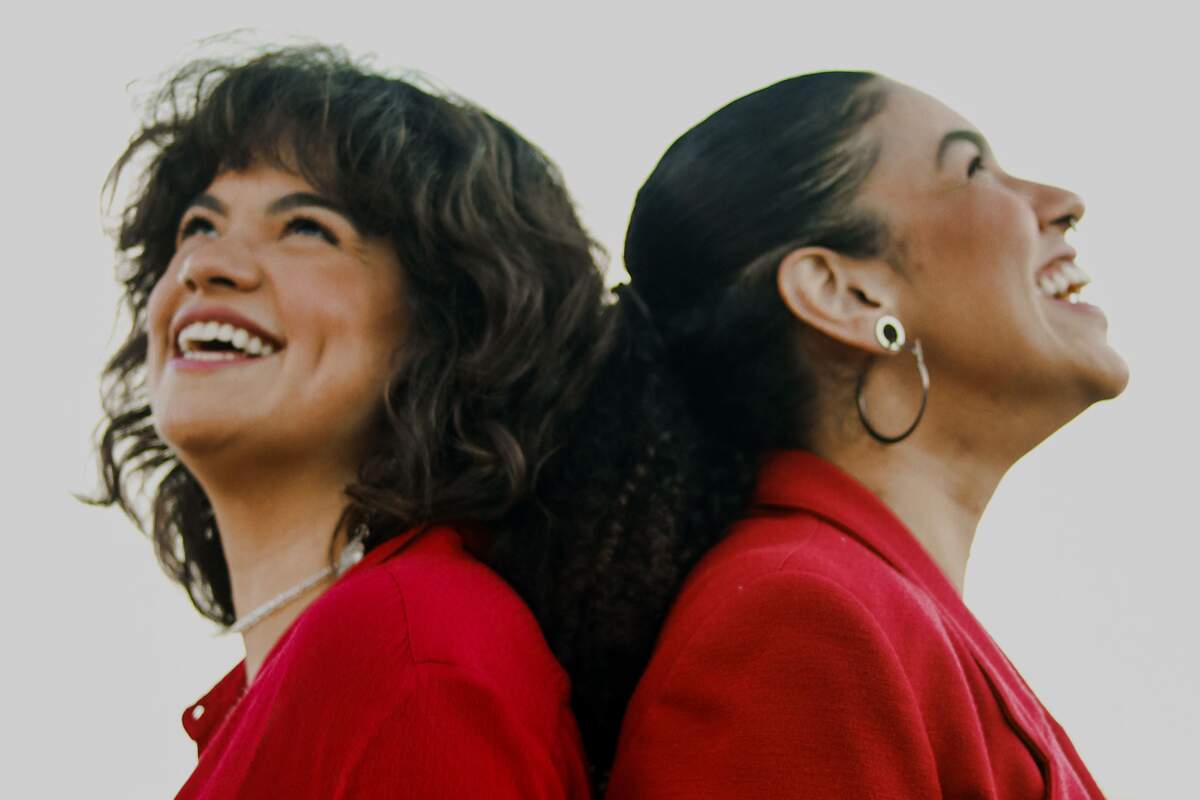 A imagem é uma fotografia de duas mulheres, do ombro para cima, de costas uma para a outra, ambas estão sorrindo, olhando para cima, vestidas com blusa vermelha e cabelos castanhos. A da esquerda está com cabelo solto e a da direita está com cabelo preso. O fundo é branco.