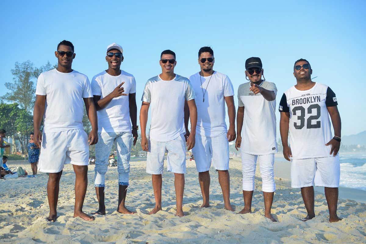 A foto mostra seis homens na praia, em pé, vestidos de branco e olhando diretamente para a frente. Todos usam óculos escuros e estão felizes. 