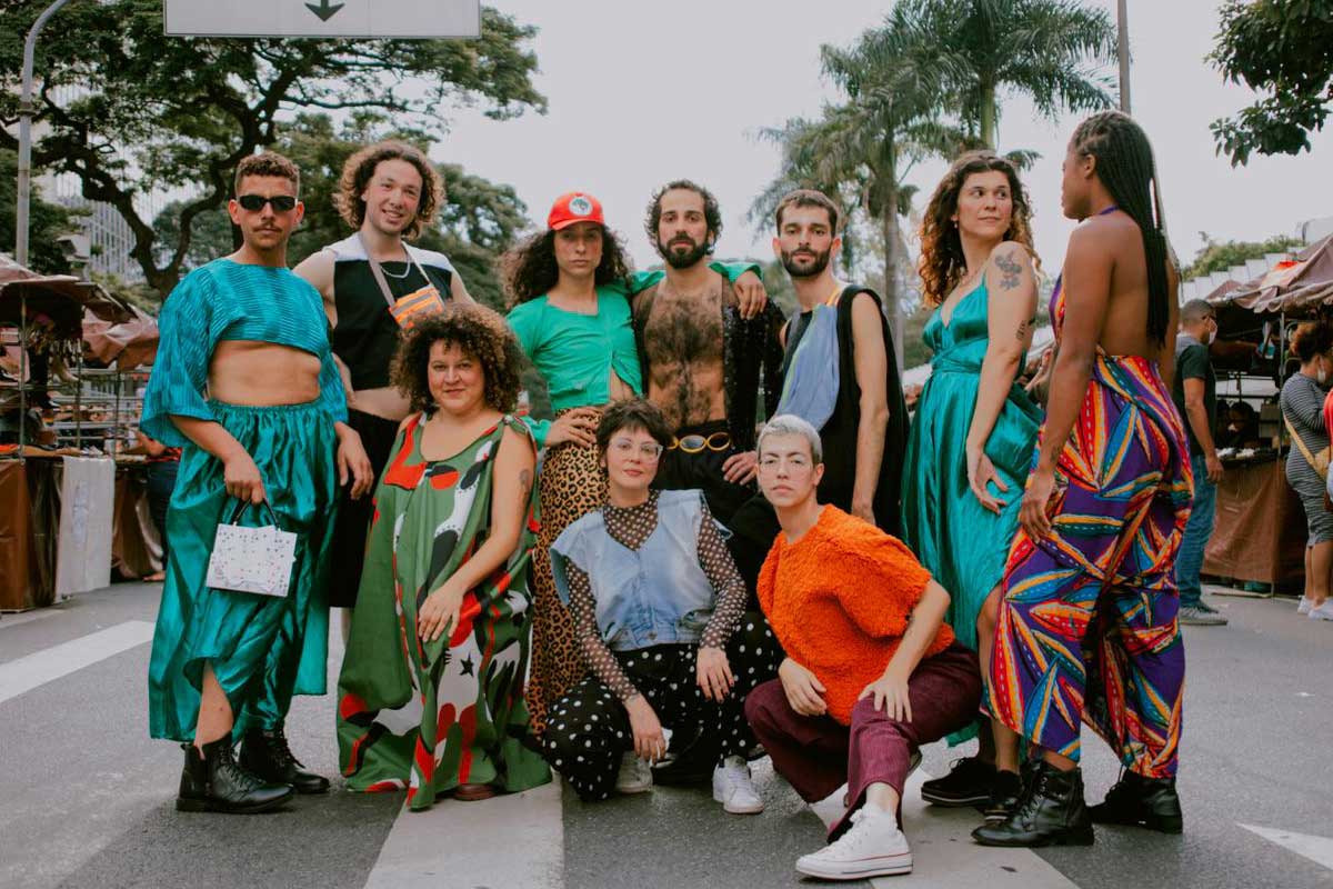 A foto mostra dez pessoas em uma feira na rua fazendo pose para a câmera. Homens e mulheres usam roupas criadas dela artista Isabela Solo. 