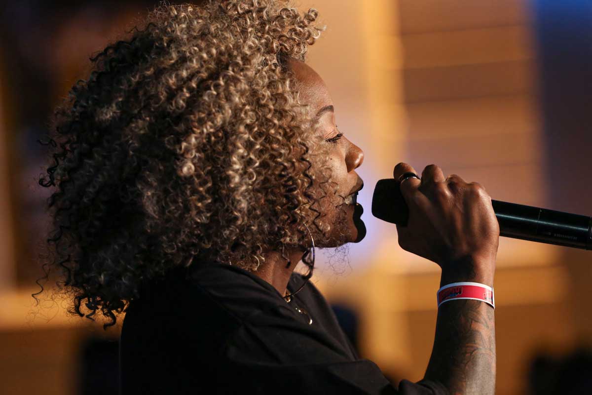 A foto mostra o perfil do rosto de uma mulher negra cantando. Com cabelos loiros e cacheados, ela usa uma camisa preta, argola dourada como brinco e um anel no dedo indicador, na mão que segura o microfone.