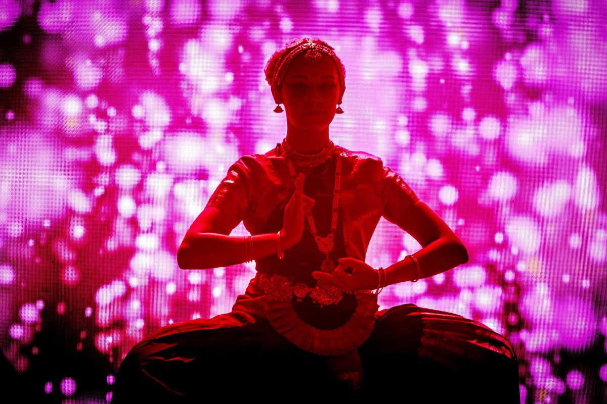 A imagem é toda em tons de rosa e há uma mulher ao centro, com trajes indianos. A pessoa está sentada sobre as próprias pernas e os braços estão em pose de meditação. A mulher usa vários acessórios como cinto, pulseiras, colares, brincos e uma tiara na cabeça.