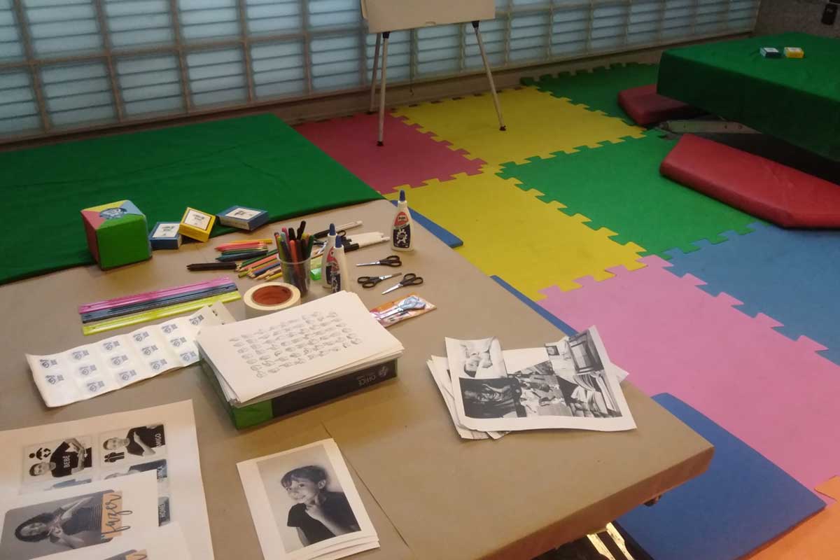 A foto mostra uma mesa com fotografias, folhas de papel, tesouras, réguas, canetas, cola e caixinhas, A mesa tem um forro verde e bege e ao fundo tem um tatame colorido no chão. À direita tem uma outra mesa e almofadas vermelhas no chão