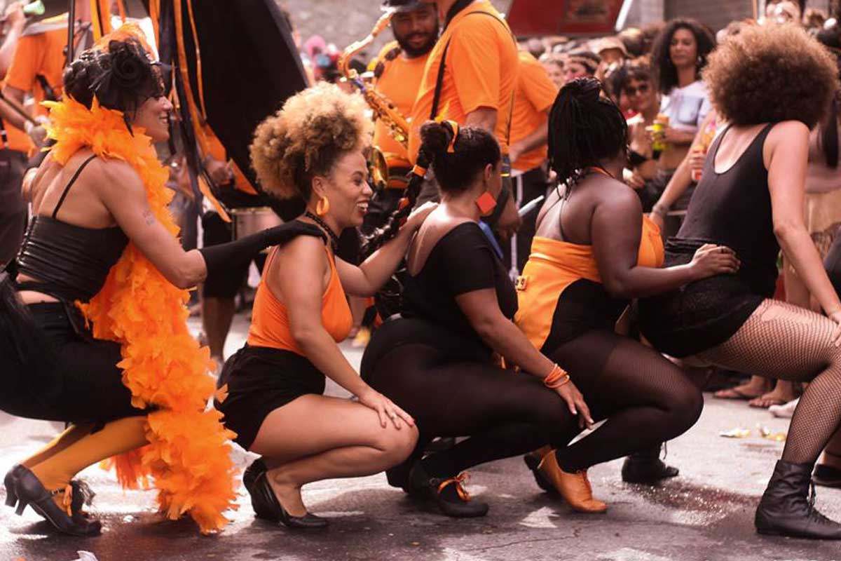 A foto mostra cinco mulheres em fila, agachadas, dançando. Elas usam roupas das cores preta e laranja, representando o bloco. Ao fundo, há homens com instrumentos musicais como saxofones, e também o público no entorno assistindo a apresentação.