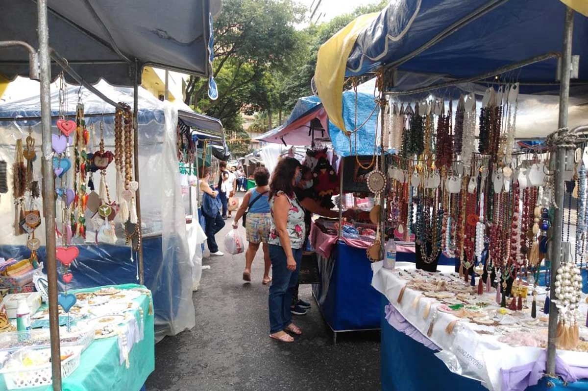 A foto mostra uma feira comercial de rua. Há barraquinhas nas laterais vendendo colares e outros acessórios e no meio as pessoas transitam e conferem as mercadorias.