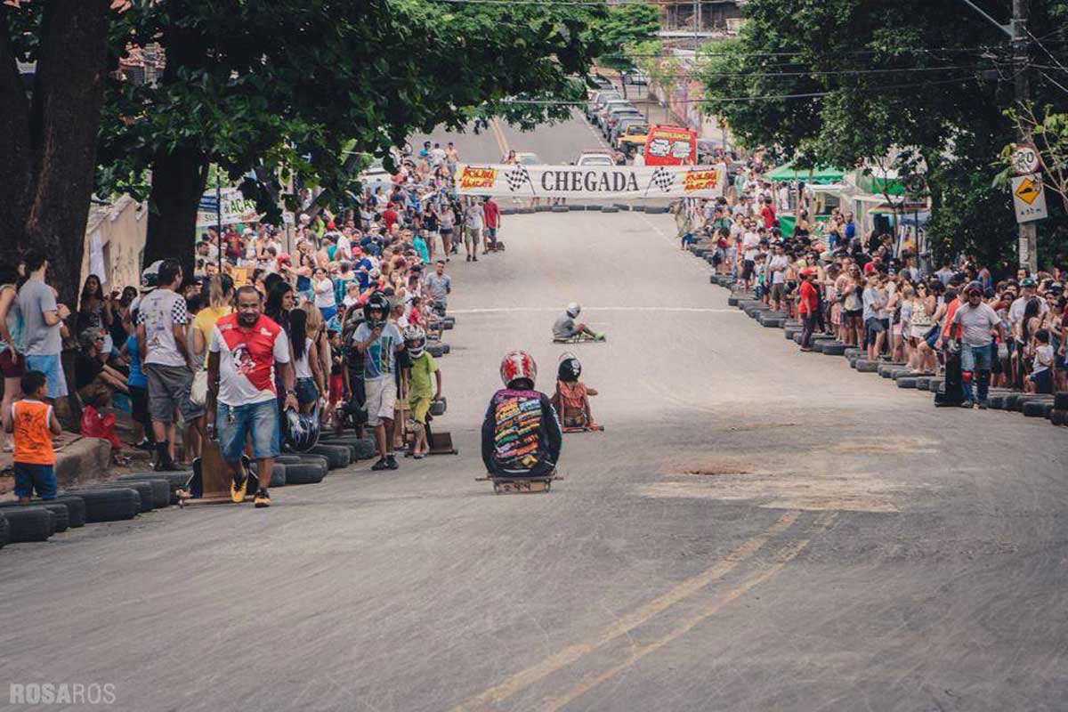 A foto mostra uma rua íngreme onde há jovens usando capacetes e andando de rolimã rumo a uma faixa com o escrito “Chegada”. Nas calçadas, o público assiste à disputa.