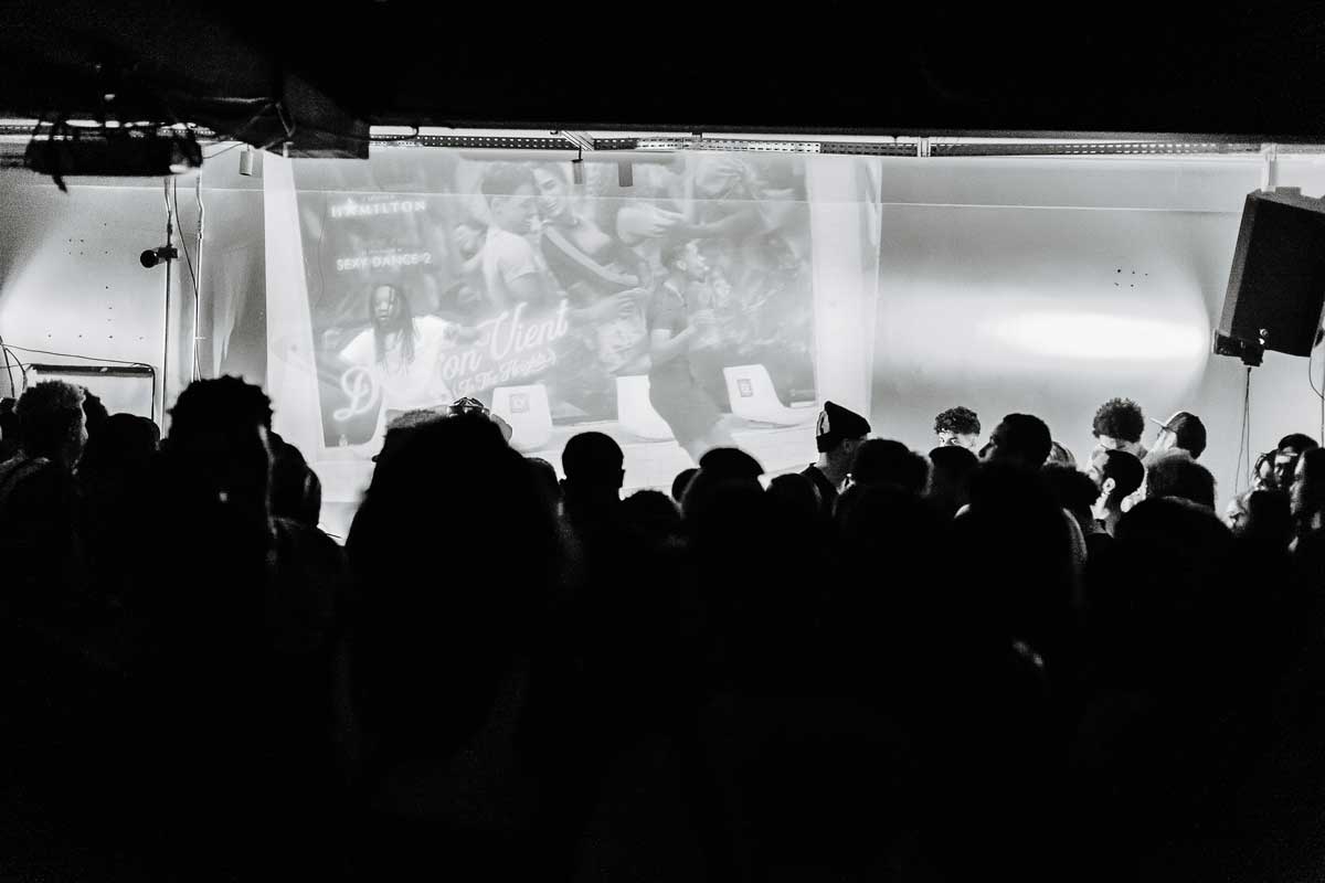  A foto em preto e branco mostra várias pessoas de costas em uma plateia, assistindo à apresentação de dois artistas. No palco, o artista à esquerda está dançando, ele tem dreads e usa uma camisa branca. O da direita, está de perfil, cantando, e usa blusa e short em cores escuras.