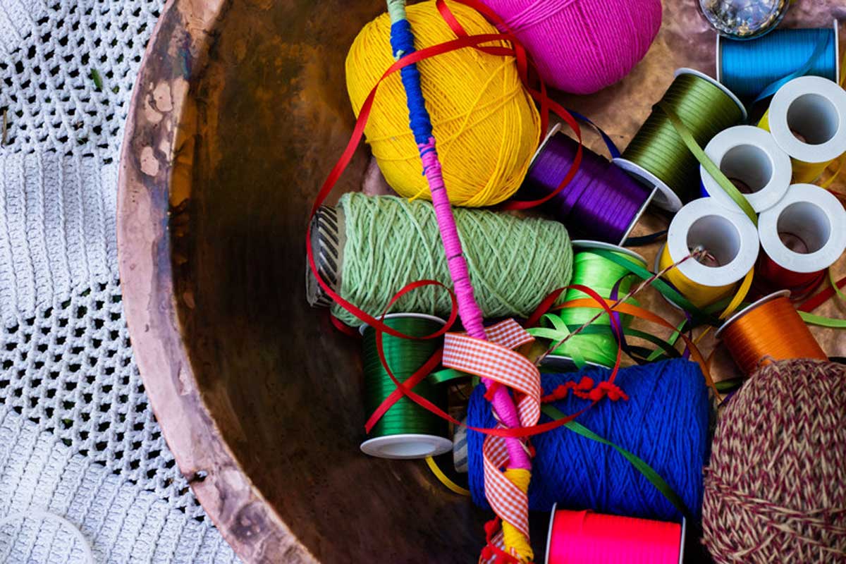 A foto mostra uma cesta com barbantes coloridos, novelos de lã, carreteis de fitilho e fitas de cetim.