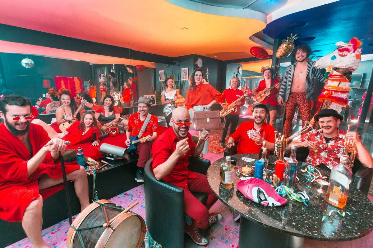 A imagem mostra várias pessoas sorrindo em um ambiente de festa. Há homens e mulheres e suas roupas são vermelhas. Algumas pessoas seguram instrumentos musicais e outras jogam baralho sentadas em uma mesa. No ambiente há confetes, balões e serpentina.	