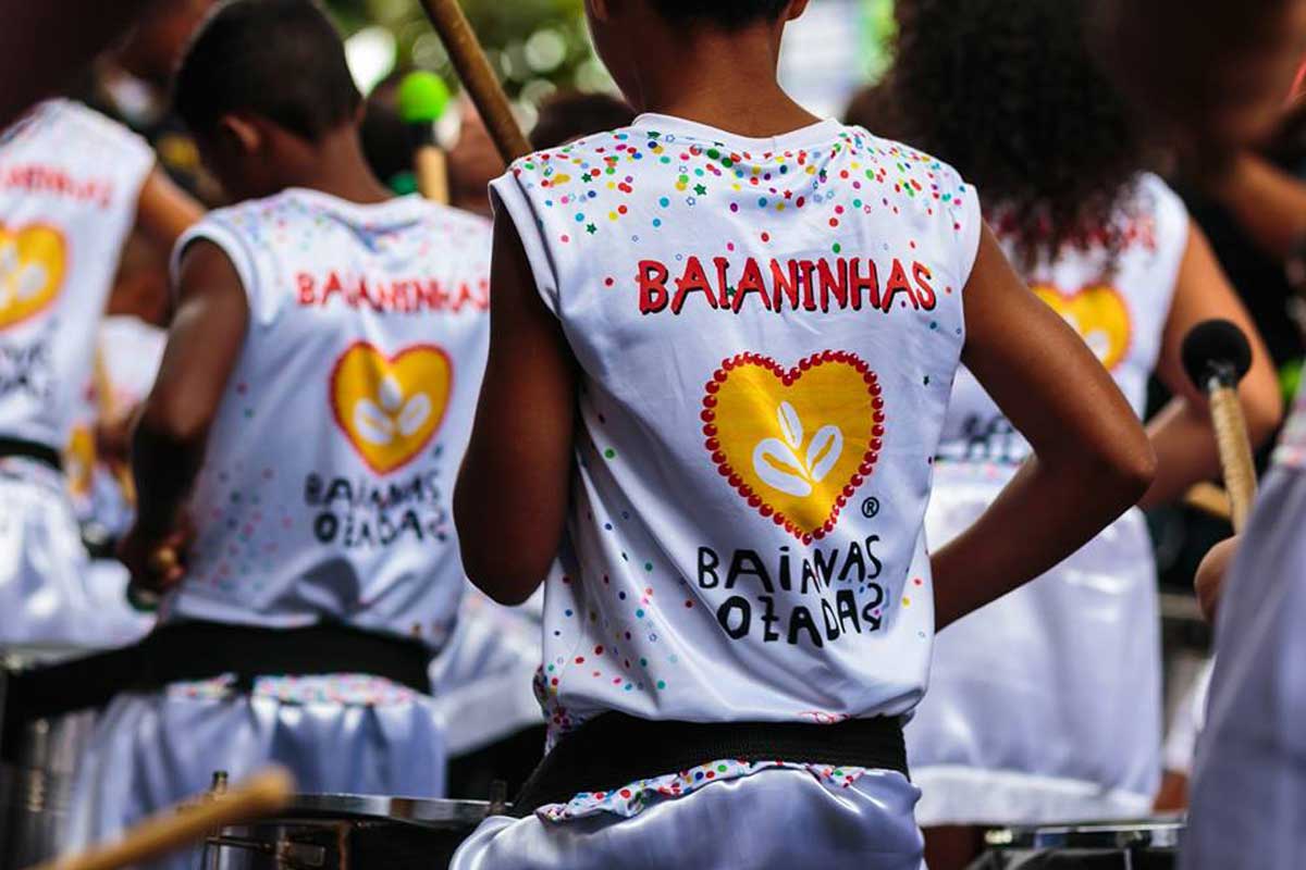 A foto mostra crianças de costas em um bloco de carnaval com abadás brancos. Na blusa das crianças há desenhos de confetes e estrelas e o escrito “Baianinhas, Baianas Ozadas”.