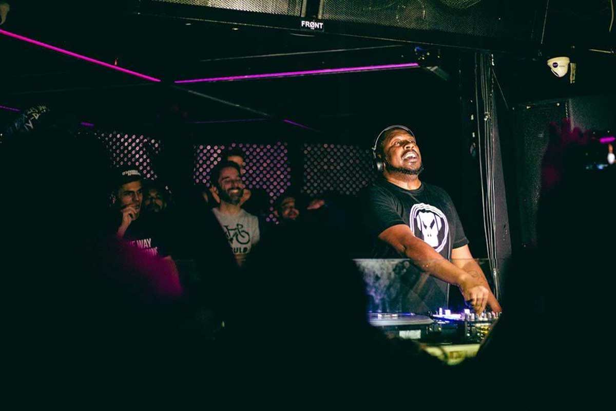 A foto mostra o DJ tocando em um ambiente fechado com luzes cor de rosa. Ele tem barba, usa um fone de ouvido e camisa preta. Em seu entorno, as pessoas dançam e se divertem.