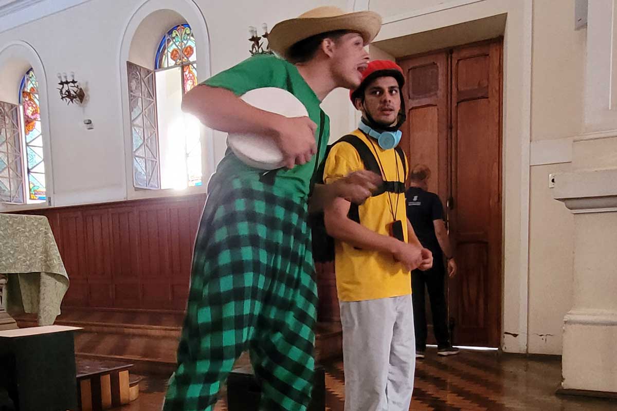 A foto mostra dois homens em uma igreja, ambos em pé, o da esquerda com um macacão verde xadrez, com uma blusa também verde por baixo. Ele segura um círculo branco na mão. O da direita usa blusa amarela, calça clara. Eles estão lado a lado e não estão olhando para a câmera