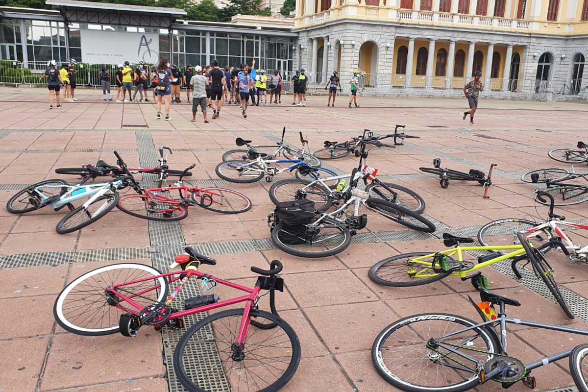 A foto mostra uma praça com várias bicicletas deitadas no chão. Ao fundo, há um aglomerado de pessoas com roupas de ciclistas como bermudas, capacetes e outros equipamentos de segurança.