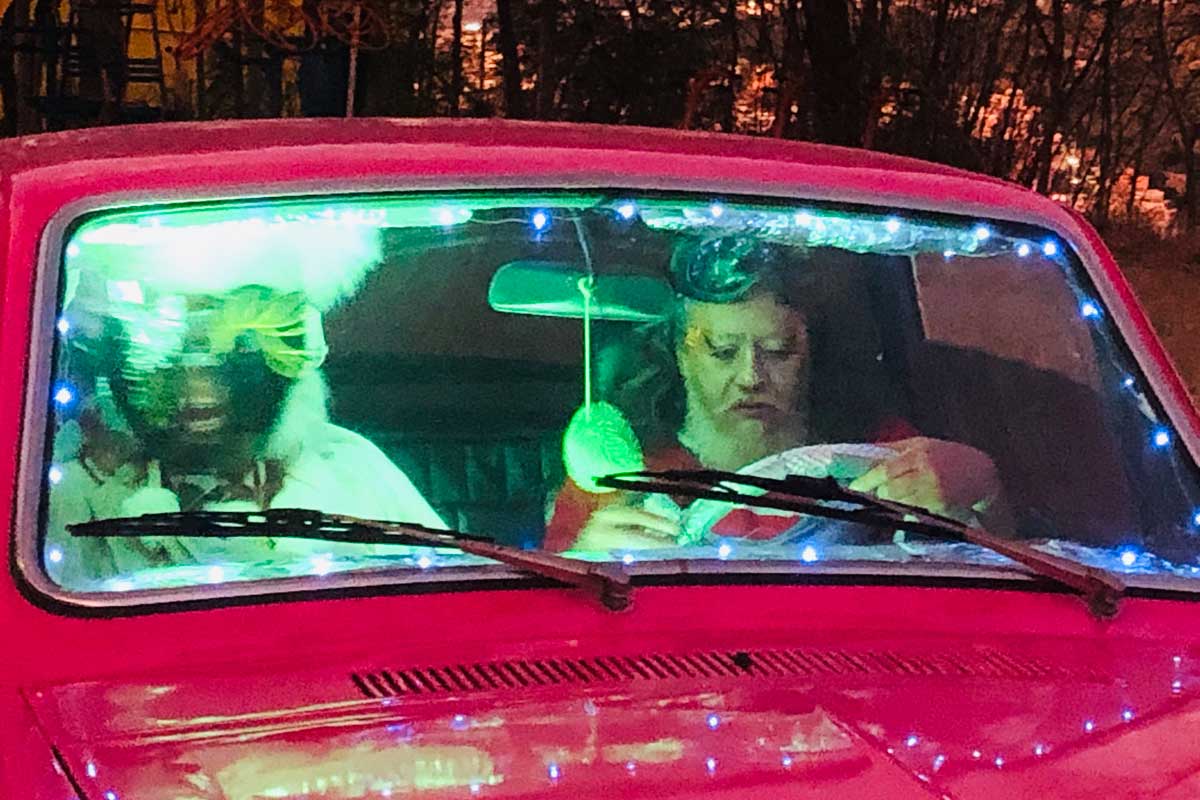 A foto mostra duas pessoas dentro de um carro rosa. Elas estão sentadas nos bancos da frente. No volante, há um homem de barba branca segurando o volante branco. Ao lado a pessoa está em movimento.