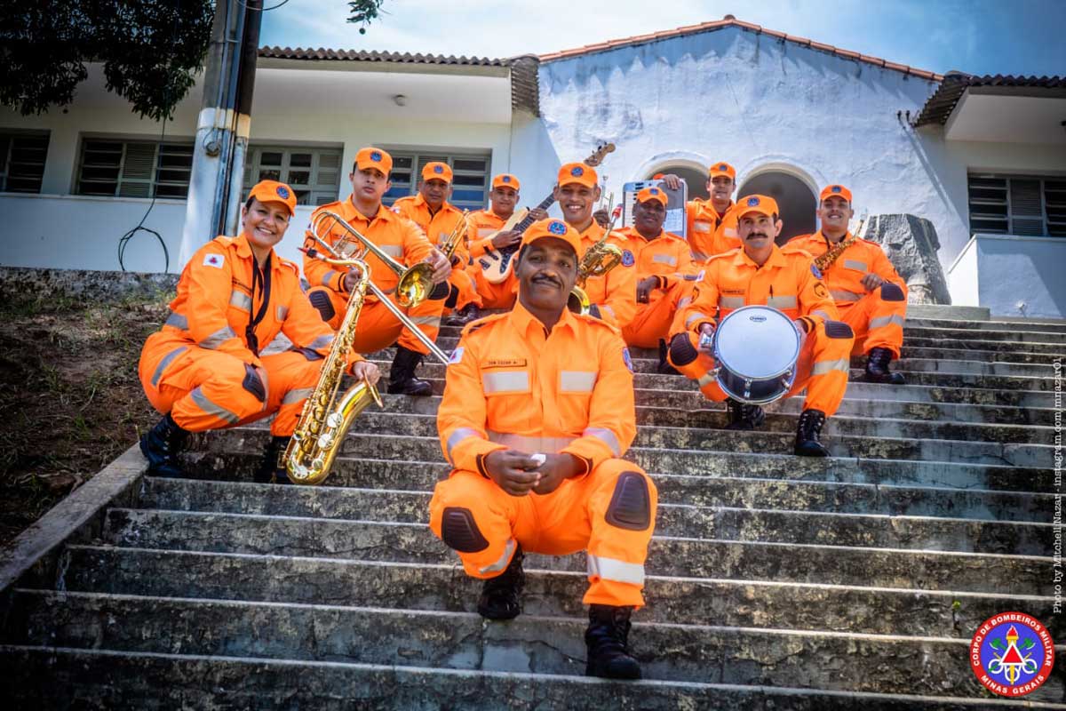 A foto mostra integrantes da Orquestra CMBIOS sentados em uma escada com seus instrumentos. Eles vestem uniforme composto por um macacão na cor laranja com detalhes em prata, boné laranja e coturno preto.