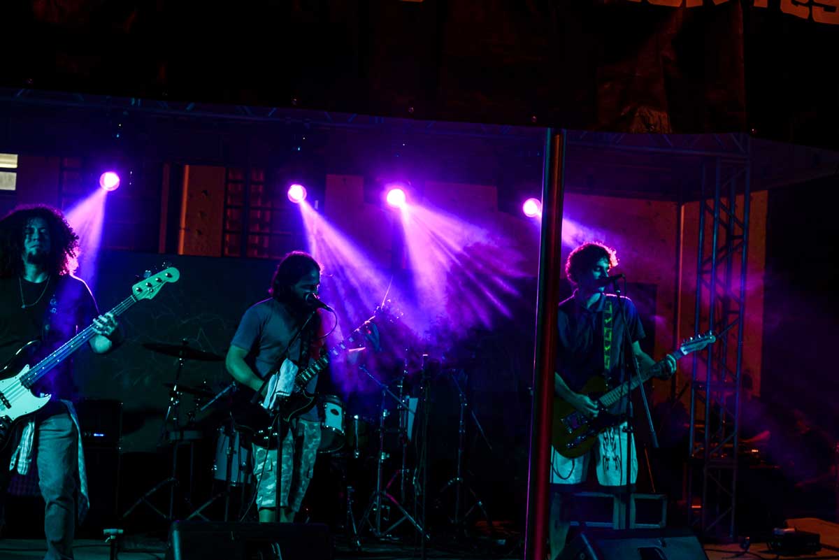 A foto mostra os integrantes da banda em um palco, à esquerda o baixista - que veste preto e tem cabelos longos encaracolados; ao centro e à direita, dois guitarristas. A luz que está sobre os artistas é verde e ao fundo do palco as luzes são roxas