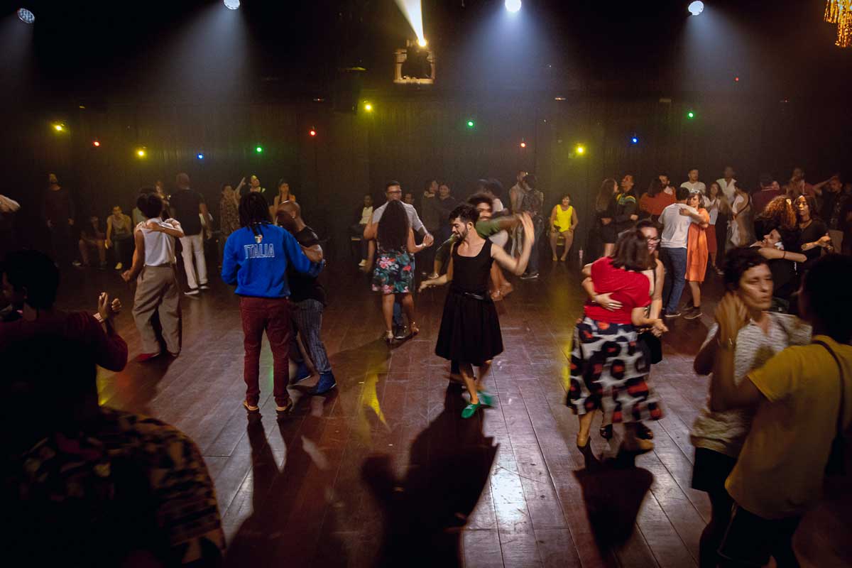  A foto mostra um salão de festas, com chão de madeira, com diversos casais dançando, no teto e ao fundo tem holofotes e luzes coloridas nas cores azul, vermelho, amarelo e verde