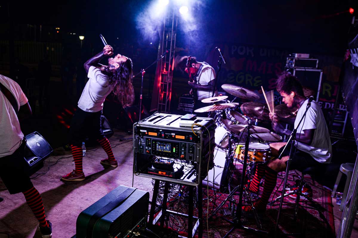 A foto mostra os quatro integrantes da banda em cima do palco, todos usam blusas brancas, meias xadrez preto e vermelho e aparecem de lado. À direita tem o baterista, ao fundo o baixista e vocalista e à direita o guitarrista. 