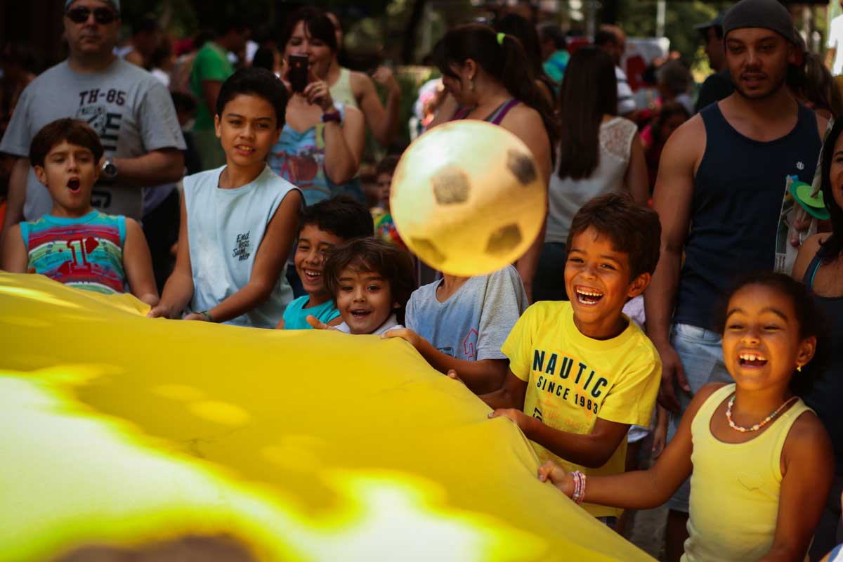 A foto mostra um grupo de crianças segurando um tecido amarelo com uma bola quicando sobre ele. As crianças estão sorrindo e olhando para a bola. Ao fundo tem diversas pessoas desfocadas. 