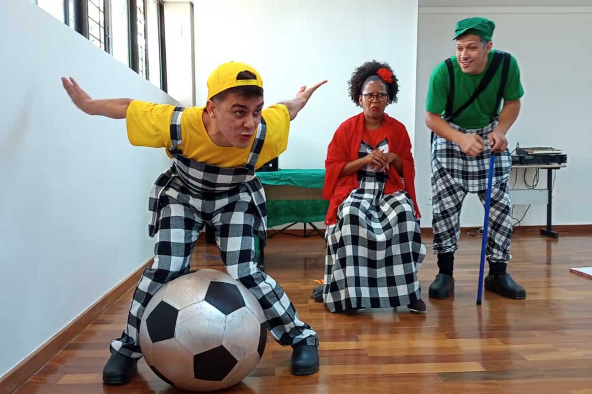 A foto mostra três adultos numa sala de chão de madeira. O da esquerda é um homem e tem uma bola entre os joelhos; no meio tem uma mulher, sentada com um lenço vermelho sobre o ombro; ao fundo, à direita, tem um homem com boné verde e bengala. Todos usam roupas xadrez