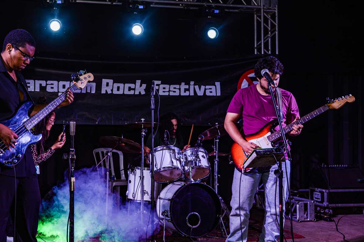A foto mostra três integrantes da banda num palco, à esquerda o baixista, que está de preto, usa óculos e olha para o instrumento. Ao centro e ao fundo tem o baterista e à direita o guitarrista, que toca uma guitarra vermelha e branco, olha para baixo e usa blusa roxa. 