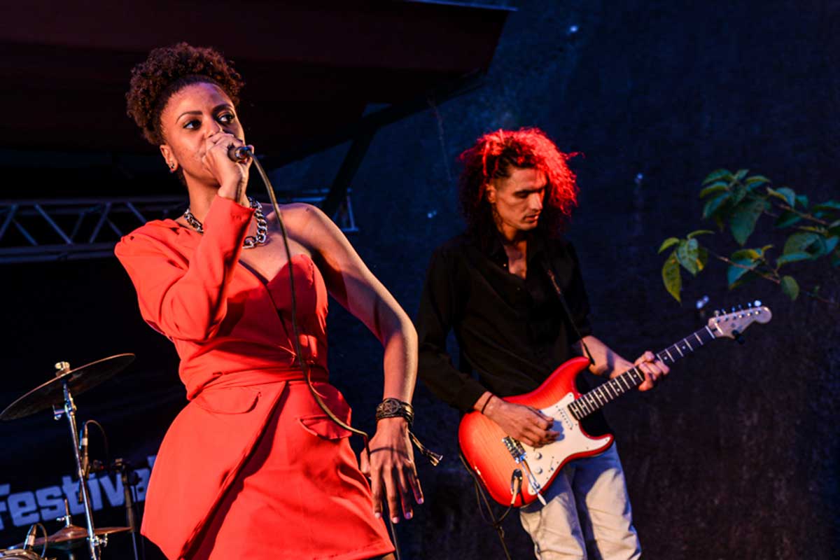 A foto mostra uma mulher, negra, com o cabelo preso, vestida com um vestido vermelho. Ela é vocalista e segura o microfone com a mão direita. Ao lado dela, à direita, aparece um homem, tocando uma guitarra vermelha e branca. No canto esquerdo aparece parte de uma bateria