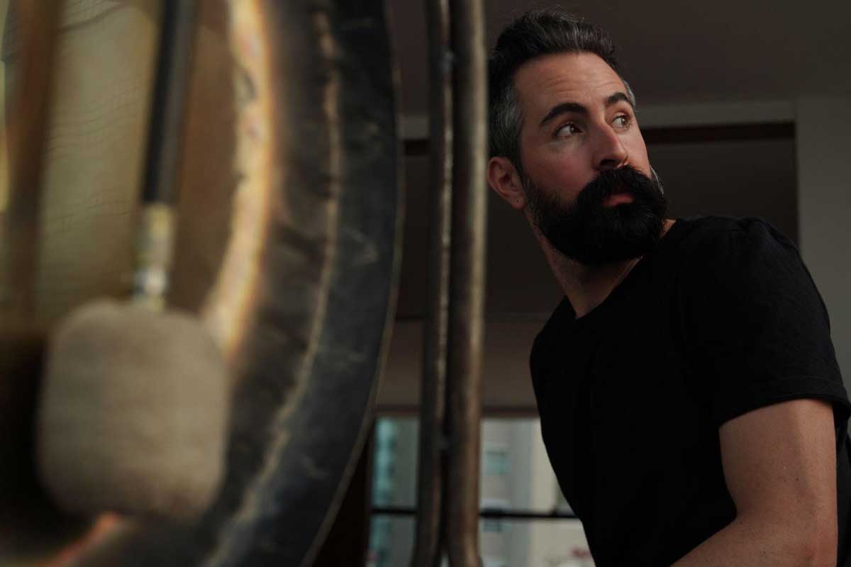 Fotografia de Renato Moura olhando por trás do ombro esquerdo. Ele veste blusa preta. No primeiro plano há um gongo antigo em desfoque.