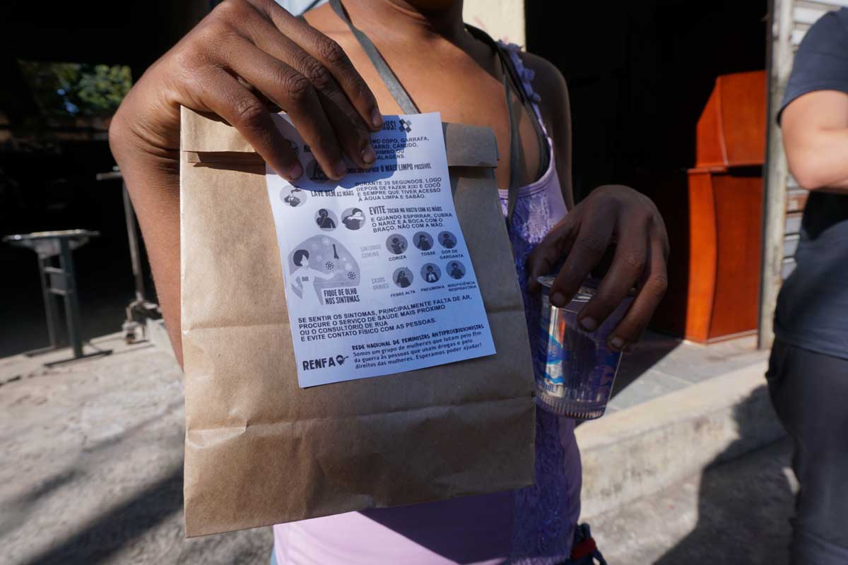 A foto mostra uma pessoa segurando um pacote pardo, um panfleto da Rede Nacional de Feministas Antiproibicionistas e um copo de água.