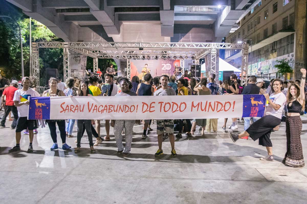 A foto mostra sete adultos, em pé, com uma faixa horizontal escrita "Sarau Vagabundo! De todo mundo!". Eles estão em um evento, embaixo de um viaduto. Ao fundo é tem um palco, sacadas de prédios e diversas pessoas de costas. 