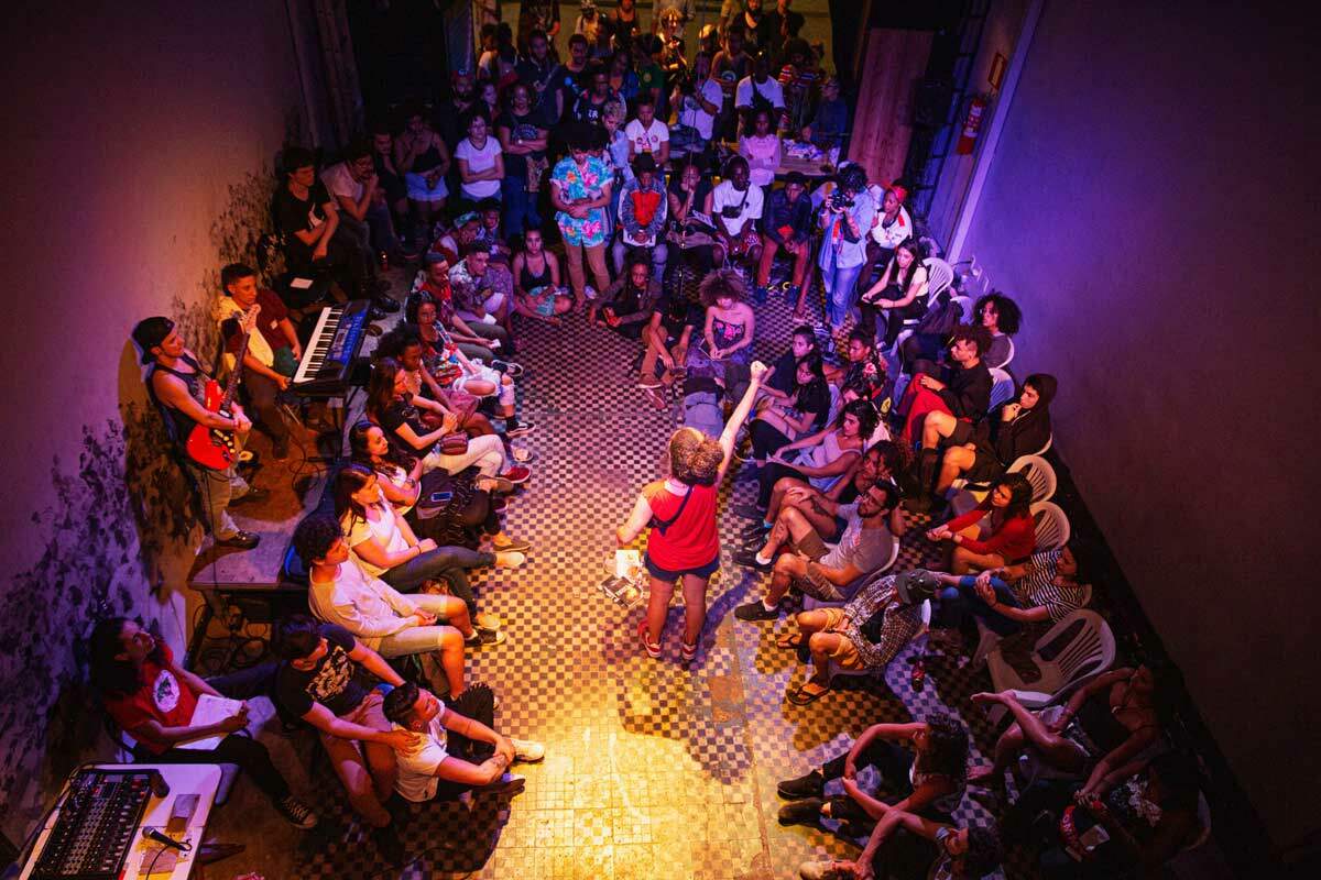 A foto mostra uma mulher de costas, com o braço direito erguido, no centro de uma roda de pessoas jovens sentadas. Eles estão em um ambiente fechado, de chão quadriculado e as luzes do ambiente são amarelas, azuis e rosas.