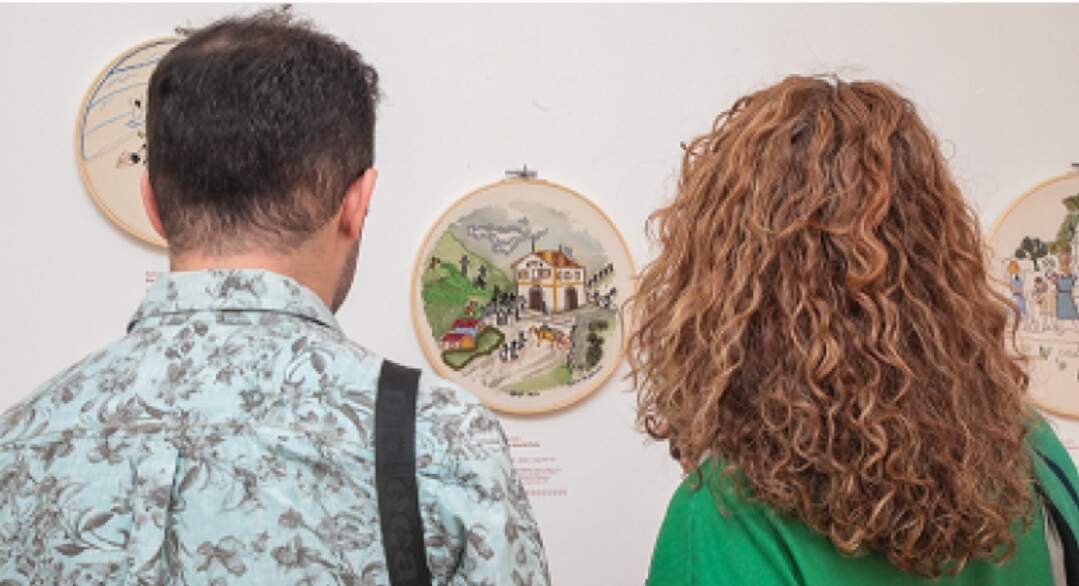 Um homem e uma mulher estão de costas para câmera observando um bordado na parede. O homem branco está usando camisa azul florida e a mulher com cabelos loiros e cacheados está usando blusa verde. 